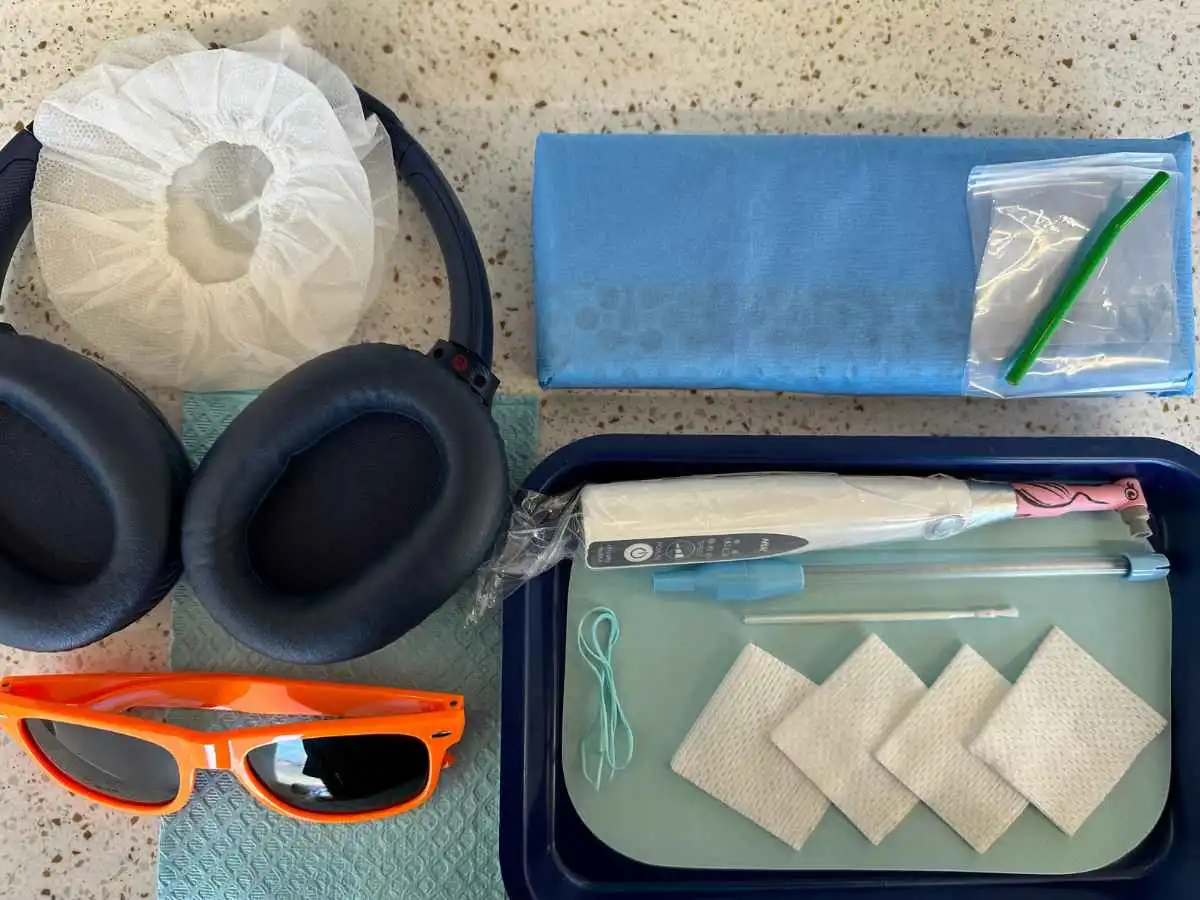 This image displays a collection of tools and products used by Poppy Kids Dental. The items are neatly arranged on a surface and include a pair of noise-canceling headphones with a hairnet stretched over the earpieces, a blue microfiber cleaning cloth, a small ziplock bag containing a green apparatus, and an open blue plastic case. Inside the case, there are a pair of orange sunglasses, a pink electronic thermometer in a protective sheath, and several adhesive bandages. The arrangement suggests these items are prepared for a specific, organized activity, for dental treatment.