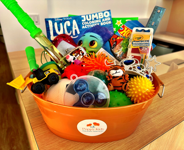 A colorful toy basket labeled "Poppy Kids" filled with a variety of children's items, including a "Disney Pixar Luca" coloring book, toy vehicles, a plush tiger, squishy sensory balls, a bubble wand, a ruler, and washable markers.